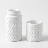 A white textured bottle with a matching small cup or lid piece, both featuring a raised honeycomb pattern, photographed on a clean white background.