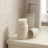 A white textured ceramic toothbrush holder filled with white cotton swabs, shown beside its matching lid on a bathroom counter with a soap dispenser and tiled wall in the background.