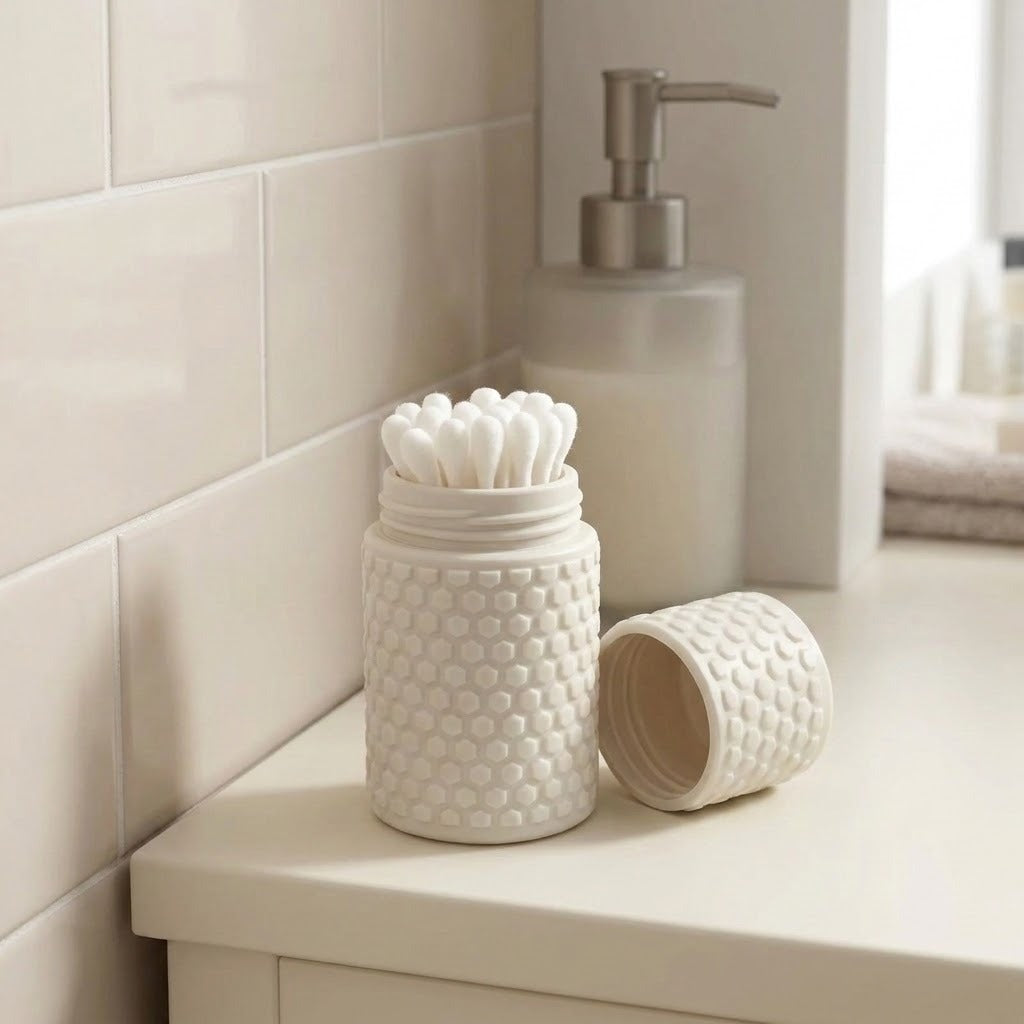 A white textured ceramic toothbrush holder filled with white cotton swabs, shown beside its matching lid on a bathroom counter with a soap dispenser and tiled wall in the background.