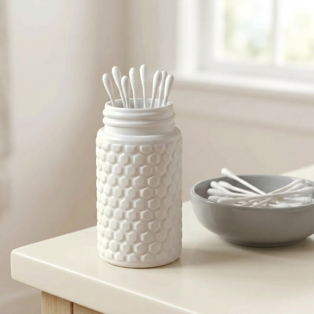 A white textured jar with cotton swab stands on a light tabletop beside a gray bowl of cotton swabs, with a softly blurred indoor background.