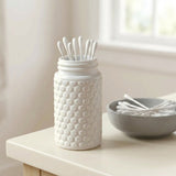A white textured jar with cotton swab stands on a light tabletop beside a gray bowl of cotton swabs, with a softly blurred indoor background.
