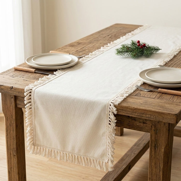 An off-white cotton table runner with fringe tassels laid on a rustic wooden dining table, styled with plates, cutlery, and a small green pine sprig with red berries.