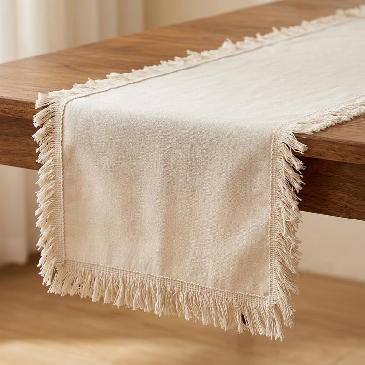 A cream-colored woven table runner with fringed edges draped over a wooden table in a warm indoor setting.
