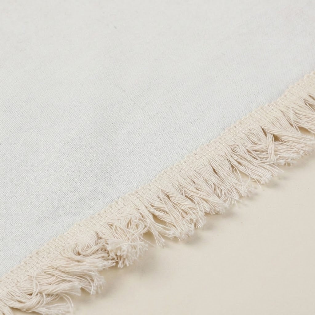 Close-up of a cream-colored woven textile with a fringed edge, shown against a matching light neutral background.