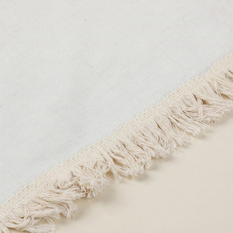Close-up of a cream-colored woven textile with a fringed edge, shown against a matching light neutral background.