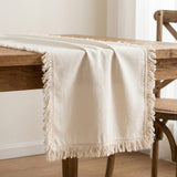 An off-white woven table runner with fringed edges draped over a rustic wooden dining table, with a wooden chair visible in the softly lit background.