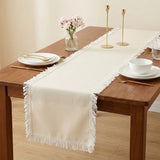 An off-white woven table runner with fringe trim laid down the center of a wooden dining table, styled with white plates, gold cutlery, and a small vase of pink flowers.