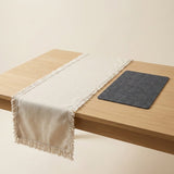 A light beige woven table runner with fringe trim draped over a light wood table, alongside a dark gray rectangular placemat on a warm cream background.