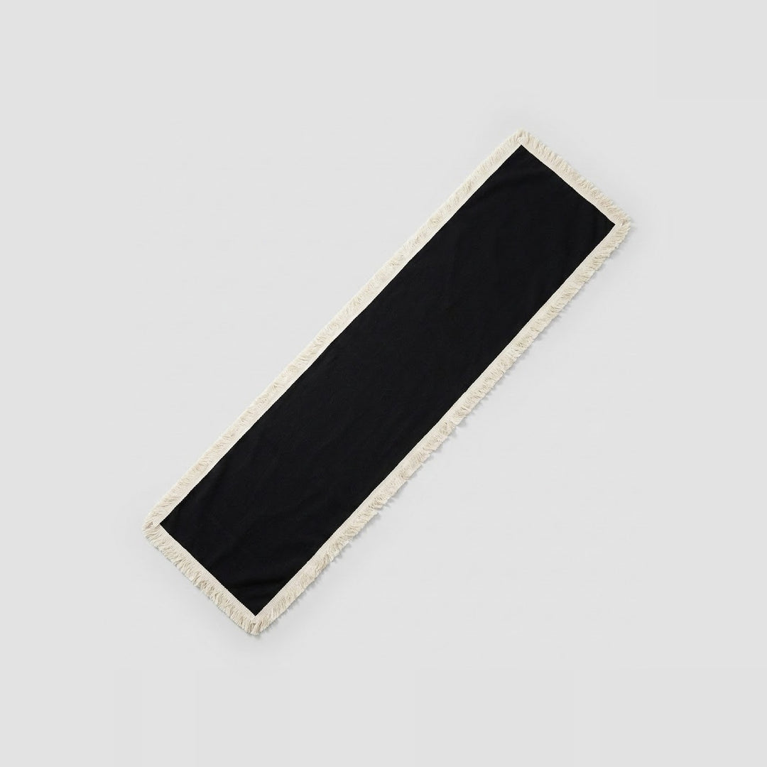 A long rectangular black scarf with a cream-colored fringed border, shown diagonally on a light gray background.
