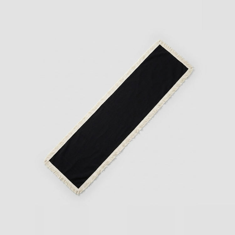 A long rectangular black scarf with a cream-colored fringed border, shown diagonally on a light gray background.