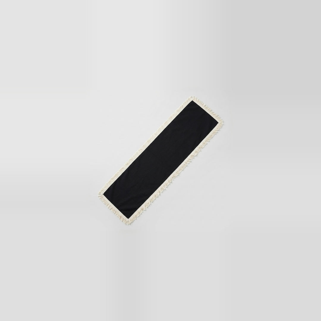 A long, narrow black rectangular mat or runner with a light beige fringed edge, shown diagonally on a light gray gradient background.