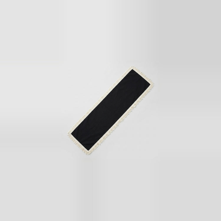 A long, narrow black rectangular mat or runner with a light beige fringed edge, shown diagonally on a light gray gradient background.