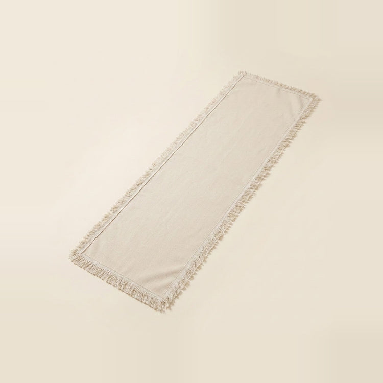 A long rectangular fringed textile runner in a warm off-white or cream tone, photographed on a plain light beige background.