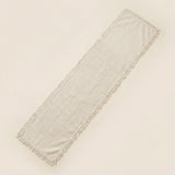 A long rectangular fringed textile runner in a light warm off-white or cream tone, photographed on a plain light beige background.