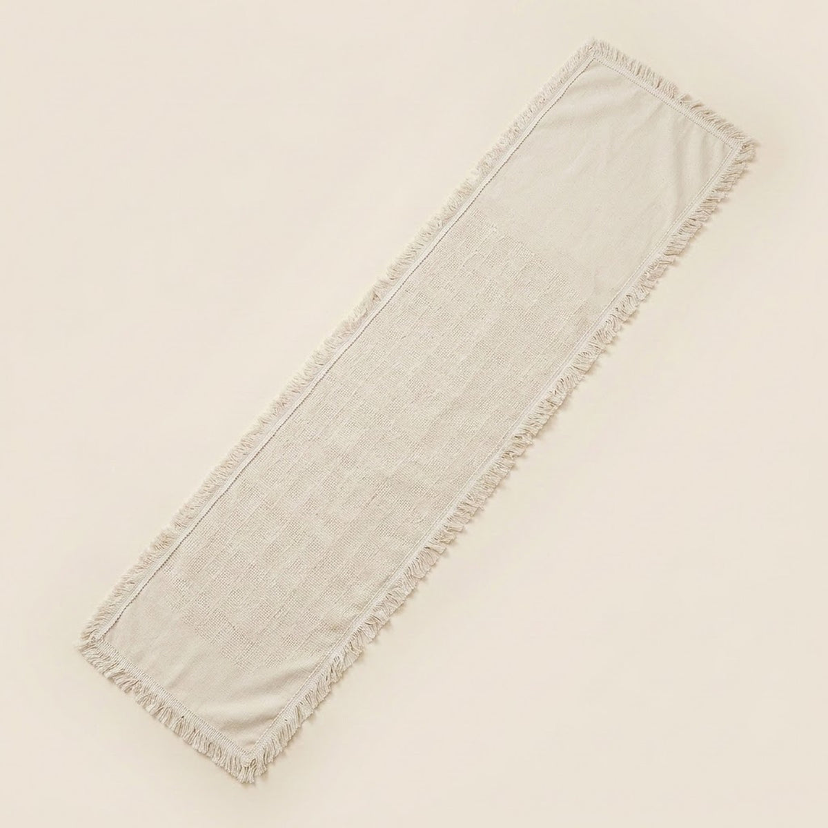 A long rectangular fringed textile runner in a light warm off-white or cream tone, photographed on a plain light beige background.