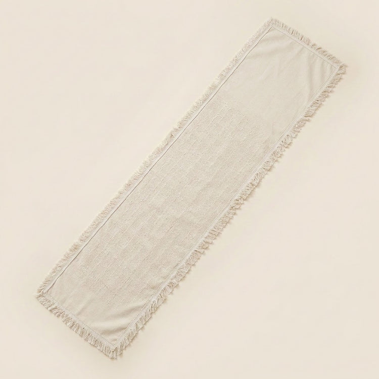 A long rectangular fringed textile runner in a light warm off-white or cream tone, photographed on a plain light beige background.