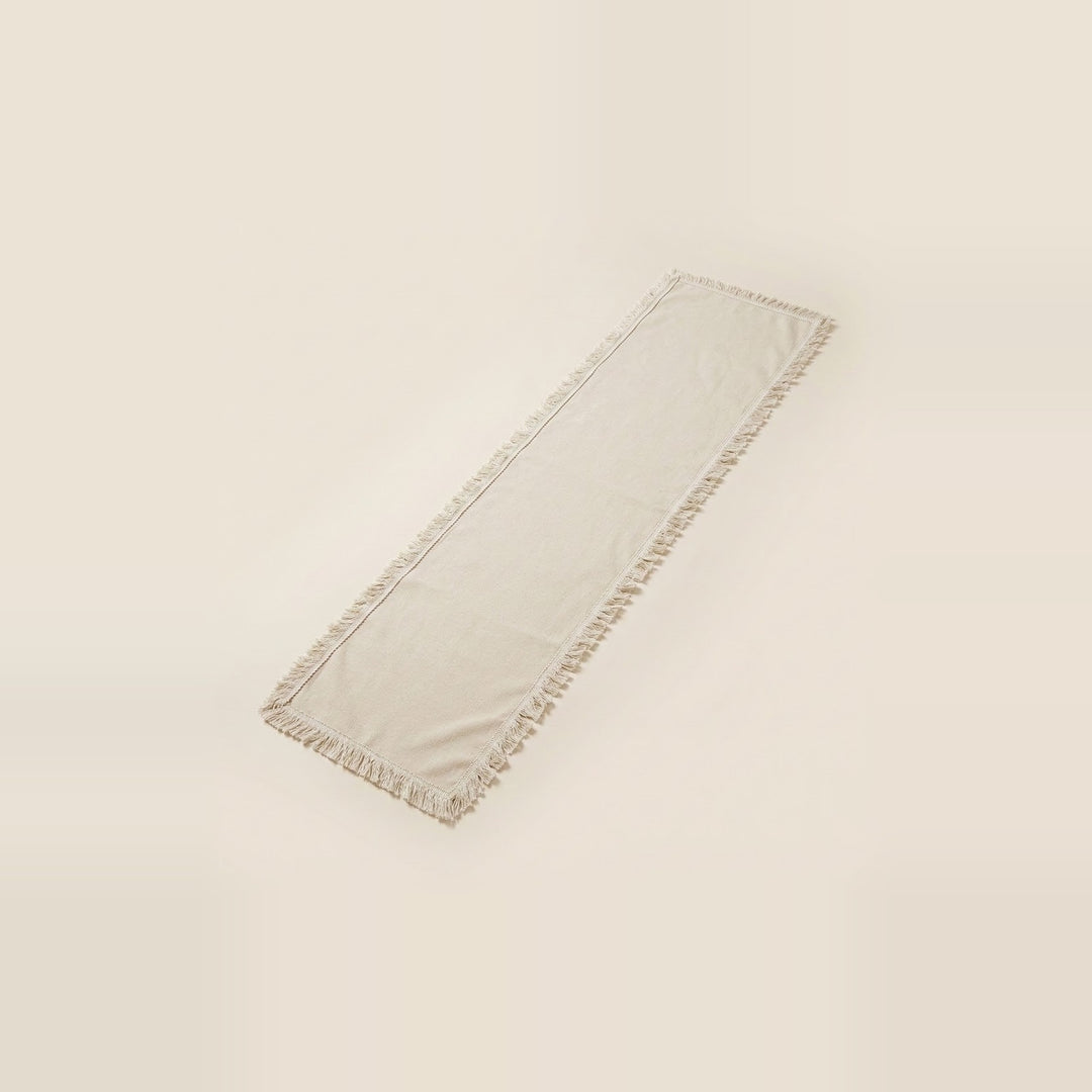 A long rectangular cream-colored table runner with fringed edges, shown on a solid light beige background.