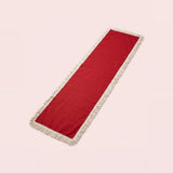 A long rectangular red runner with cream fringe on both ends, photographed diagonally on a solid pale pink background.