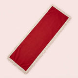 A long rectangular red runner with cream fringe on both ends, laid diagonally on a solid pale pink background.