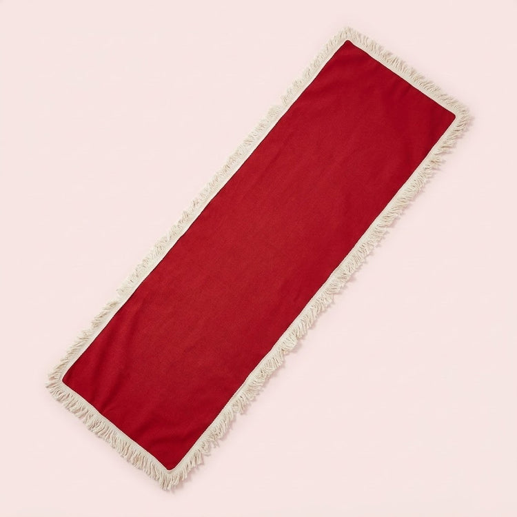 A long rectangular red runner with cream fringe on both ends, laid diagonally on a solid pale pink background.