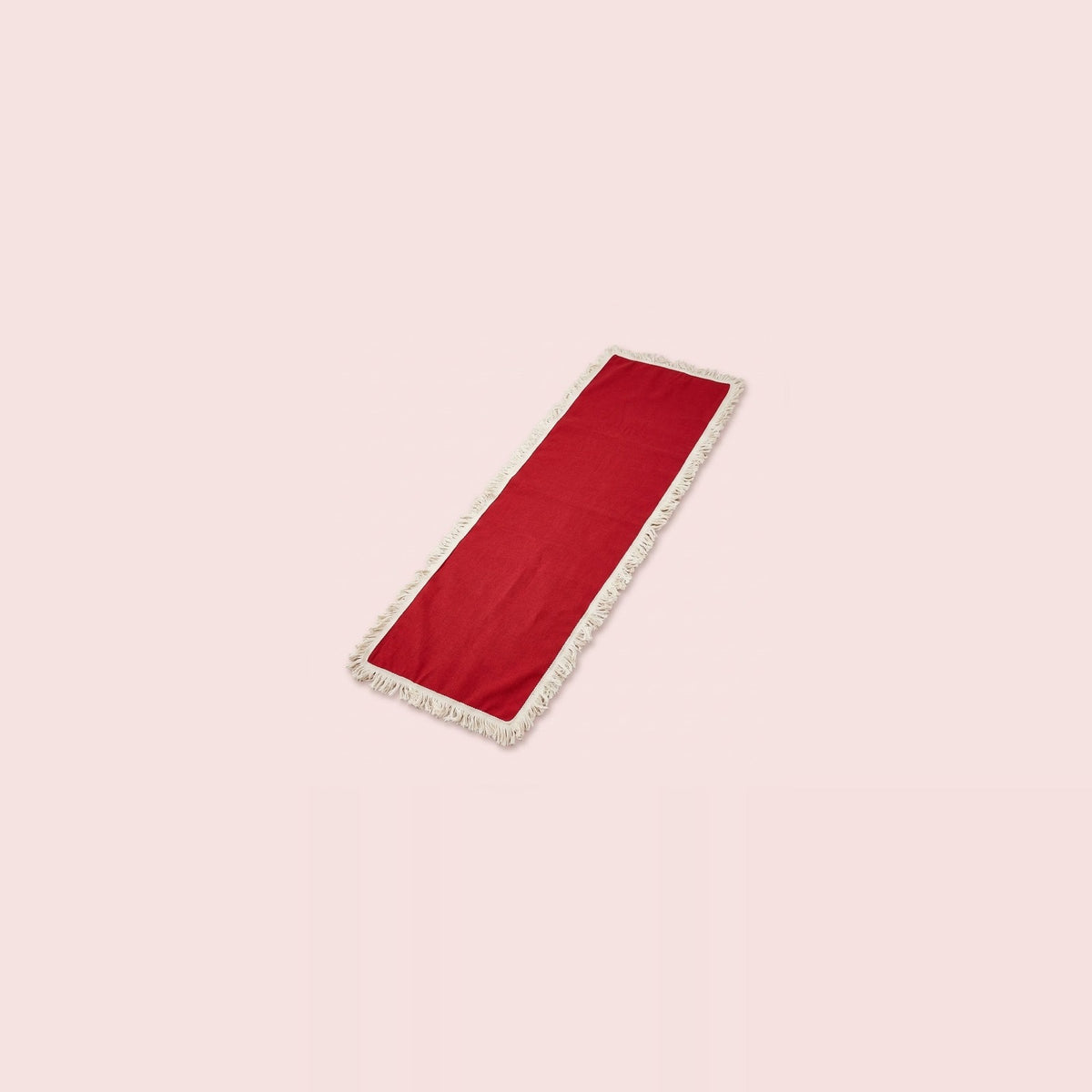 A long rectangular red runner with white fringe trim on both ends, displayed diagonally on a solid pale pink background.