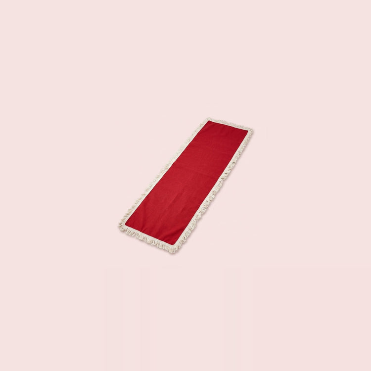 A long rectangular red runner with white fringe trim on both ends, displayed diagonally on a solid pale pink background.