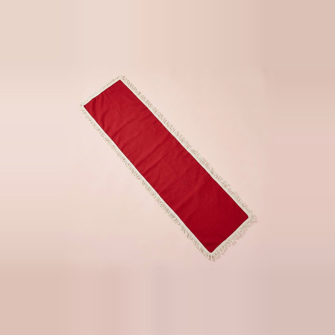 A long rectangular red runner with white fringe trim, laid diagonally on a solid pale pink background.