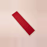 A long rectangular red runner with white fringe trim, laid diagonally on a solid pale pink background.