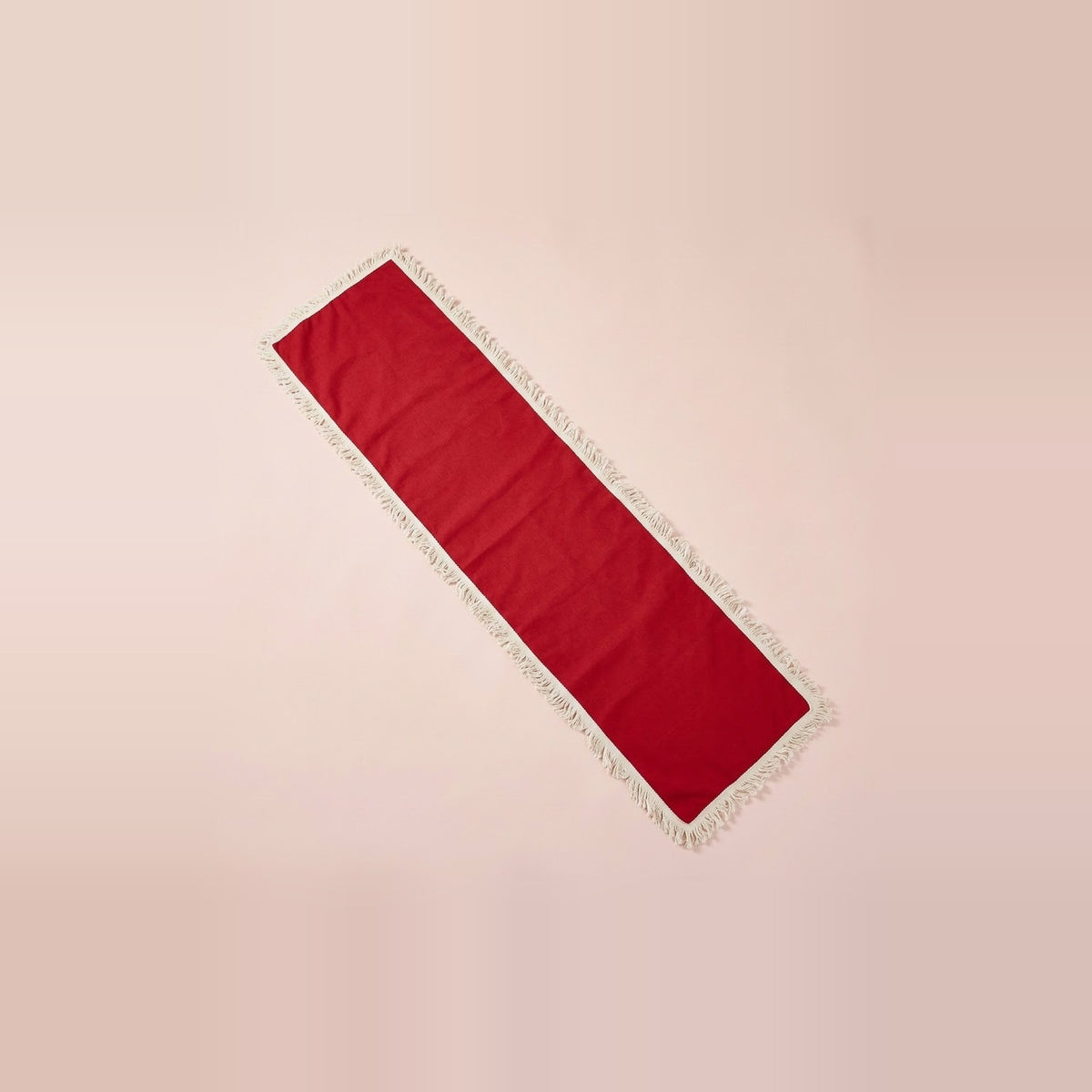 A long rectangular red runner with white fringe trim, laid diagonally on a solid pale pink background.