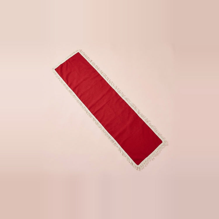 A long rectangular red runner with white fringe trim, laid diagonally on a solid pale pink background.