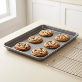 A dark gray metal baking sheet holding six chocolate chip cookies on a light beige countertop, with a wire cooling rack beside it and a softly blurred kitchen background.