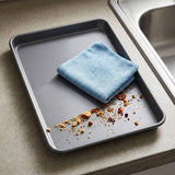 A gray rectangular metal baking pan sits on a beige countertop next to a stainless steel sink, with a folded light blue microfiber cloth and scattered crumbs inside the pan.