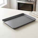 A dark gray rectangular baking sheet resting on a light beige countertop in a kitchen setting, with a stainless steel oven and a folded white towel in the background.