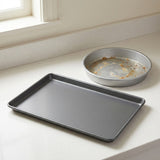 A dark gray rectangular baking sheet and a round silver metal pie pan resting on a light beige countertop near a window.