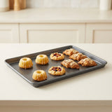 A dark gray rectangular baking sheet holding six baked pastries, including two ring-shaped pastries with glaze and four almond pastries with fruit filling, set on a light beige countertop in a kitchen.