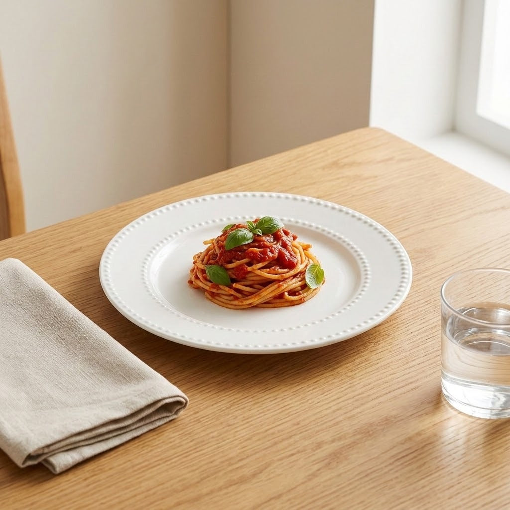A white ceramic plate with a decorative beaded rim sits on a light wood table, styled with a small serving of spaghetti with tomato sauce and basil leaves. A folded beige napkin and a glass of water are placed nearby in a bright, natural setting.