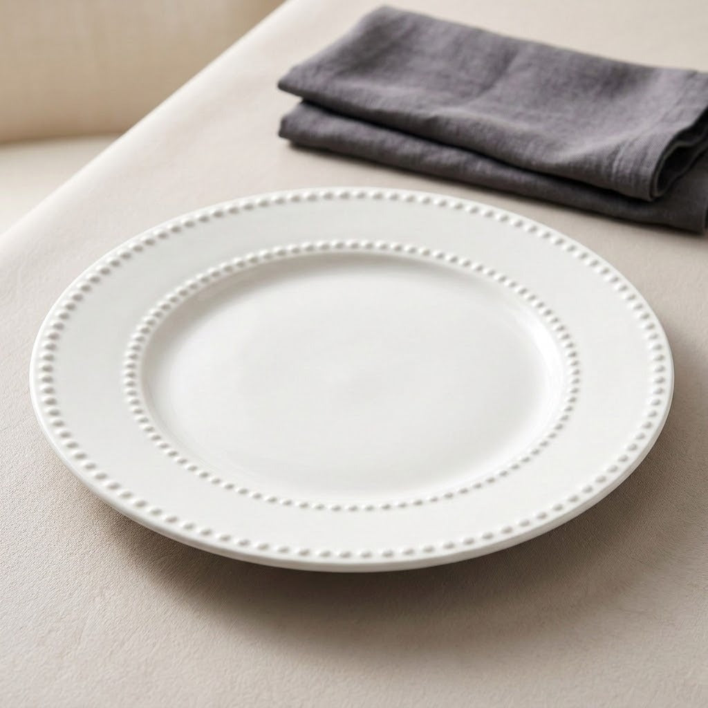 A white ceramic dinner plate with a decorative beaded border, shown on a light beige tablecloth with a folded gray napkin in the background.