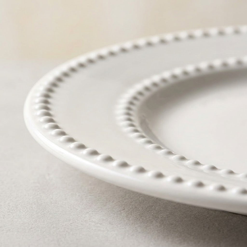 A close-up of a white ceramic plate with a decorative beaded rim, resting on a neutral gray surface.