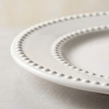 A close-up of a white ceramic plate with a decorative beaded rim, resting on a neutral gray surface.