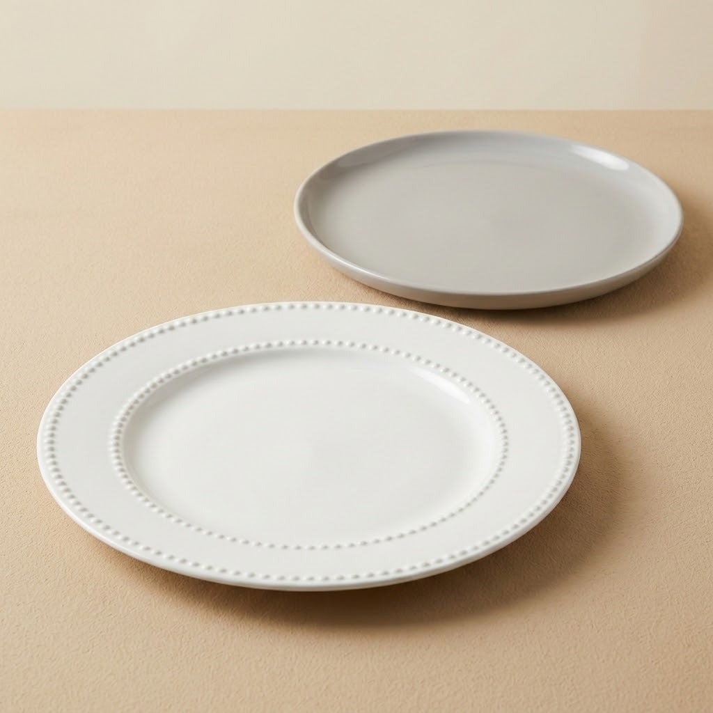 Two round ceramic plates on a beige surface, with one white plate in the foreground featuring a beaded rim and a light gray plate in the background.