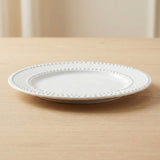 A white ceramic plate with a decorative beaded border, resting on a light wood tabletop with a softly blurred neutral background.