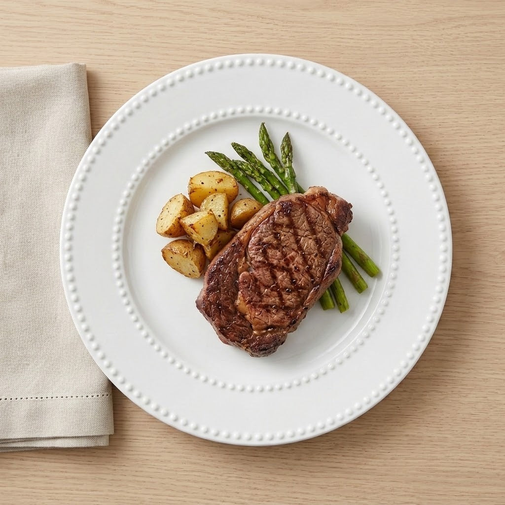 A white ceramic plate with a beaded rim, styled with a grilled steak, green asparagus, roasted potato chunks, and a folded beige napkin on a light wood tabletop.