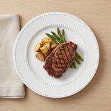 A white ceramic plate with a beaded rim, styled with a grilled steak, green asparagus, roasted potato chunks, and a folded beige napkin on a light wood tabletop.