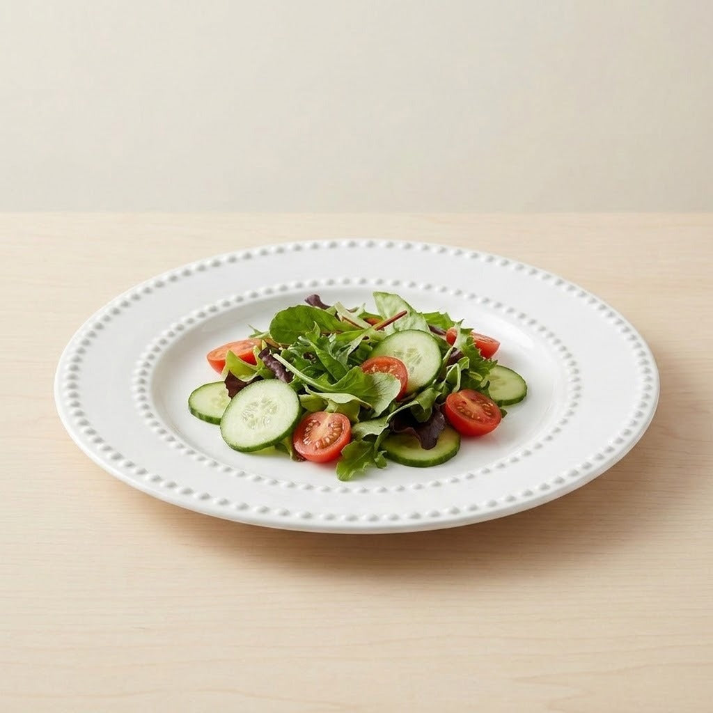 A white ceramic plate with a decorative beaded rim, holding a fresh green salad with cucumber slices and cherry tomatoes, set on a light wood tabletop against a neutral beige background.