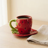 A red strawberry-shaped ceramic mug with a green handle and stem, filled with coffee and resting on a matching saucer on a wooden table next to a beige napkin.