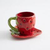 A red strawberry-themed ceramic cup with a matching saucer, featuring green leafy stems and black seed details, photographed on a clean white background.