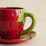 A red strawberry-shaped ceramic cup with a green handle and leaf detail, shown on a matching saucer against a light beige background.