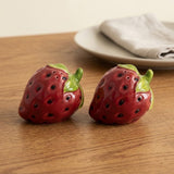 Two strawberry-shaped salt and pepper shakers with glossy red bodies, green leafy tops, and black seed details, placed on a wooden tabletop with a blurred plate and napkin in the background.