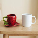 A red strawberry-themed ceramic cup with a green handle and stem sits on a matching saucer on a wooden table, next to a plain white ceramic mug.