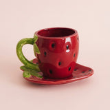 A glossy red ceramic strawberry-shaped cup with a green handle and leaf, shown on a matching saucer against a soft pink background.